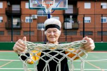 Старшая женщина в спортивной одежде и повязке смотрит в камеру, стоя с сеткой в руке на баскетбольной площадке с обручем — стоковое фото