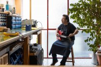 Mestre em afinação avental guitarra enquanto joga perto da mesa com vários instrumentos e ferramentas sentados na cadeira perto de janelas e vaso planta em quarto brilhante — Fotografia de Stock