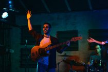 Молодой человек, играющий на бас-гитаре, выступая в легком клубе с неоновой подсветкой, машет рукой публике — стоковое фото