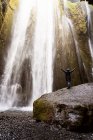 Vista posterior de un turista irreconocible con una cálida chaqueta encapuchada de pie en un cañón rocoso con una potente y pintoresca cascada Gljufrafoss con brazos extendidos durante el viaje a Islandia - foto de stock