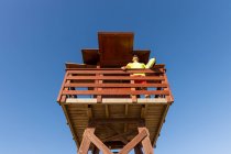 Angle bas de sauveteur courageux dans des lunettes de soleil debout sur la tour de sauvetage en bois et surveillant la sécurité sous le ciel bleu — Photo de stock