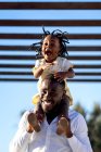 Heureuse fille afro-américaine avec des tresses sombres assises sur les épaules du père joyeux et sautant tout en s'amusant ensemble dans la rue au soleil — Photo de stock