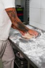 Side view of crop tattooed male baker in white polo shirt kneading dough with hands while standing at metal counter in kitchen of modern bakery — Stock Photo