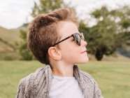 Зміст хлопчика в теплому одязі і модних сонцезахисних окулярах, що відпочивають на трав'янистому полі проти пишних зелених дерев у природі в літній день — стокове фото