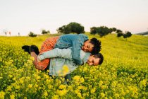 Romantische junge Mann lächelt und gibt huckepack Fahrt zu freudigen afroamerikanischen Freundin in üppig blühenden gelben Wiese in der Landschaft — Stockfoto