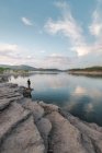 Далекий неузнаваемый человек, стоящий на грубом каменистом побережье, наслаждаясь спокойным озером в природе на фоне голубого неба и холмистой местности — стоковое фото