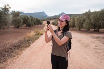 Smiling Asian female in cap taking picture in a camp of green olive tree with a modern smartphone while standing in grove — Stock Photo