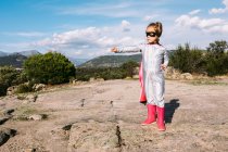 Повне тіло маленької дівчинки в костюмі супергероя, що піднімає витягнуті кулаки для показу влади, стоячи на скелястому пагорбі — стокове фото