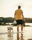 Cuerpo completo de propietario masculino anónimo con botas en las manos caminando en el agua cerca de perro corriente en el día de verano en la naturaleza - foto de stock