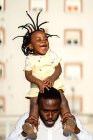 Fröhlicher afroamerikanischer Vater im Hemd, der seine kleine Tochter auf den Schultern trägt und springt, während er die Zeit zusammen auf der Straße in der Stadt im Sonnenlicht verbringt — Stockfoto