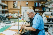 Seitenansicht des aufmerksamen Senior-Kunsthandwerkers in Schürze und Brille, der vor der Arbeit an der Druckpresse Klebebänder auf Holzplatten bindet — Stockfoto