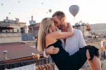 Hombre sosteniendo novia en las manos mientras está de pie con los ojos cerrados en la terraza en el fondo de globos de aire caliente a la luz del sol - foto de stock