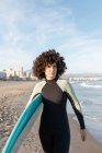Jovem surfista feminina pensativa em roupa de mergulho com prancha de surf andando olhando para longe na praia lavada pelo mar ondulante — Fotografia de Stock