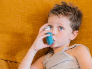 Вид збоку маленького хлопчика з астмою, який використовує інгалятор, сидячи на дивані вдома — стокове фото