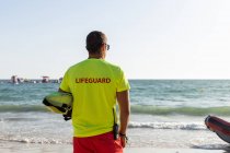 Vue arrière du sauveteur masculin anonyme en short et t-shirt et maintien de la sécurité sur le bord de mer sablonneux — Photo de stock