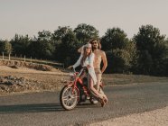 Ganzkörper-Hippie-Pärchen auf rotem Moped auf Asphaltfahrbahn bei Ausflug in die Natur mit Bäumen an Sommertagen — Stockfoto