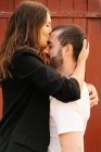Seitenansicht des romantischen jungen bärtigen Mannes in lässiger Kleidung trägt glückliche Freundin und küsst mit geschlossenen Augen in der Nähe Tür auf der Straße an sonnigen Tag — Stockfoto