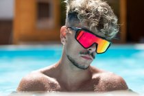 Confident shirtless man with curly hair in sunglasses in swimming pool with splashing water on sunny summer day in resort — Stock Photo