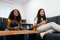 Positive amiche multirazziali sedute sul divano e che giocano ai videogiochi mentre trascorrono del tempo insieme a casa — Foto stock