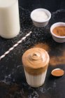 Bicchiere di delizioso caffè Dalgona con latte e condimento schiumoso posto su tavolo disordinato nero con cacao in polvere e zucchero — Foto stock