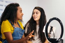 Positive female with long dark hair in casual clothes sitting looking at each other and showing at cheerful African American blogger while recording vlog on modern smartphone placed on tripod with LED ring lamp at home — Stock Photo