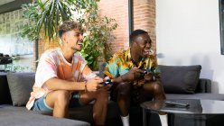 Amigos multirraciais alegres com gamepads em mãos sentadas no sofá enquanto jogam videogame juntos na sala de estar leve com planta verde — Fotografia de Stock