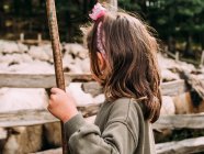 Cute little girl in casual clothes playing with wooden stick standing on grassy meadow near flock of sheep in enclosure in farmyard — Stock Photo