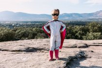 Full body of small girl in superhero costume with hands on waist standing on rocky hill — Stock Photo