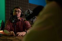 Молодой радиоведущий в клетчатой рубашке и наушниках сидит за столом с микрофоном и общается с анонимным коллегой во время записи подкаста в студии — стоковое фото