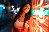 Charming female in white top looking at camera while standing near building with glowing lights in evening time on street — Stock Photo
