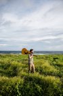 Спокійний музикант у повсякденному одязі, що стоїть з акустичною гітарою на плечі серед зеленої трави на узбережжі океану в літній час в денний час — стокове фото