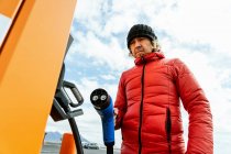 From below of thoughtful male in warm outwear and hat standing with charging power to electric vehicle in hand at gas station in sunny day — Stock Photo