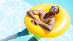Calma macho preto em calções de banho e óculos de sol banhos de sol no anel inflável amarelo na piscina com água clara no dia ensolarado de verão — Fotografia de Stock