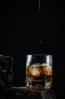 Whiskey drops falling on ice cubes served in crystal glass placed on rough surface against black background — Stock Photo