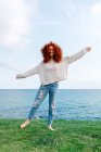 Full body of happy curly haired female outstretching arms while enjoying freedom on grassy hill on coast of sea — Stock Photo