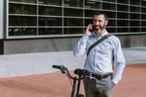 Trabajador masculino en ropa formal de pie con bicicleta y conversando por teléfono móvil en el centro - foto de stock