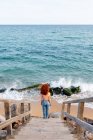 Vista trasera de ángulo alto de viajera femenina irreconocible con pelo largo de jengibre rizado de pie en la playa de arena húmeda bañada por olas salpicadas de espuma - foto de stock