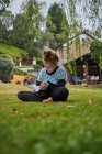Ganzkörper konzentriertes Barfußmädchen liest interessantes Buch, während es im Hinterhof auf einer Rasenfläche vor einem Wohnhaus auf dem Land sitzt — Stockfoto