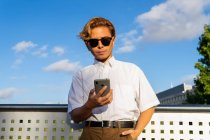 Homem jovem confiante em mensagens de texto camisa branca no celular, enquanto em pé na rua contra o céu azul no dia ensolarado — Fotografia de Stock