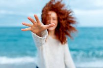 Pleasant female with long ginger curly hairstyle reaching hand to camera while standing against waving sea — Stock Photo