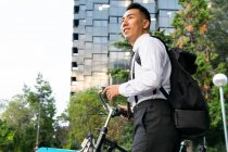 Vista lateral de soñador joven oficinista étnico masculino con mochila y bicicleta mirando hacia otro lado contra edificio urbano y árboles - foto de stock