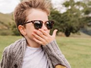 Content boy in warm clothes and trendy sunglasses resting on grassy field against lush green trees in nature on summer day — Stock Photo