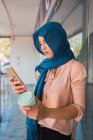 Восхитительная мусульманка в хиджабе и с кофе, чтобы пойти просматривать мобильный телефон, стоя на улице города и глядя на экран — стоковое фото