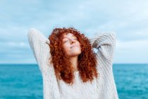Восхитительная женщина с кудрявыми длинными рыжими волосами в трикотажном свитере, стоящая с руками за головой и закрытыми глазами против синего моря — стоковое фото