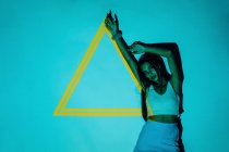Trendy millennial ethnic female with long hair and raised arm looking at camera against yellow triangle from projector light — Stock Photo