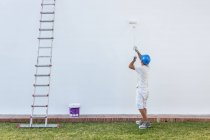 Jovem pintor irreconhecível pintando a fachada de um apartamento com um rolo com a escada ao lado — Fotografia de Stock