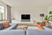 Удобный диван и кресло, расположенные рядом с телевизором против окон и дверей в гостиной современной недвижимости — стоковое фото