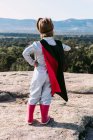 Back view of small unrecognizable girl in superhero costume with hands on waist standing on rocky hill — Stock Photo