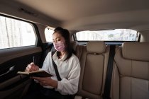 Mujer vestida formalmente con máscara protectora escribiendo en bloc de notas mientras monta en el asiento trasero del pasajero en cabina cómoda - foto de stock