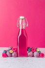 Glasflasche mit frischem Fruchtsaft umgeben von reifen Beeren auf dem Tisch serviert mit Gläsern auf rosa Hintergrund — Stockfoto